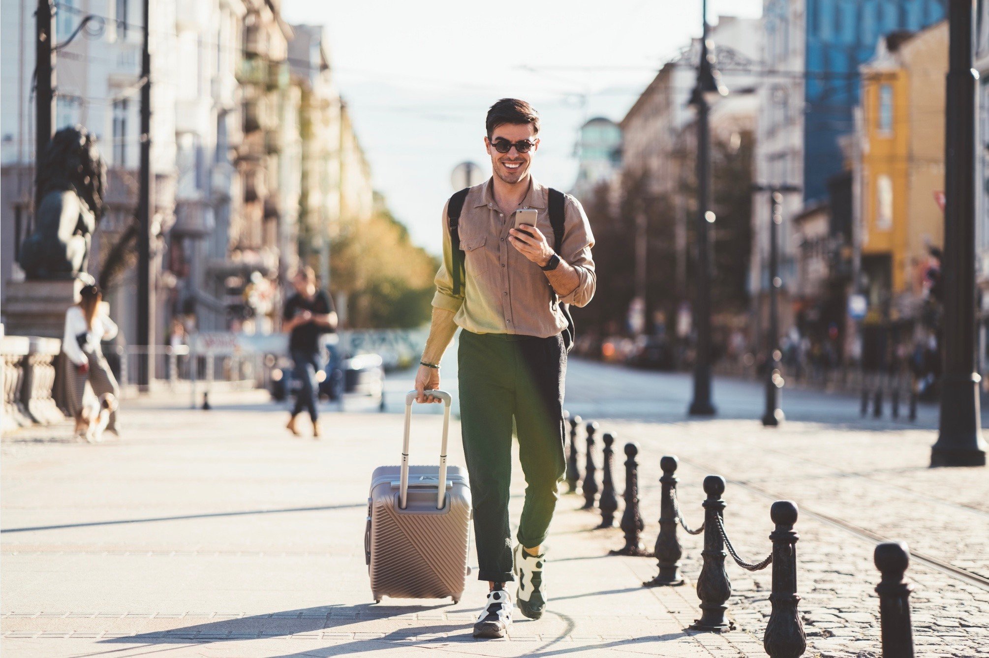 Man looking down at his phone, walking down the street with a rolling travel bag