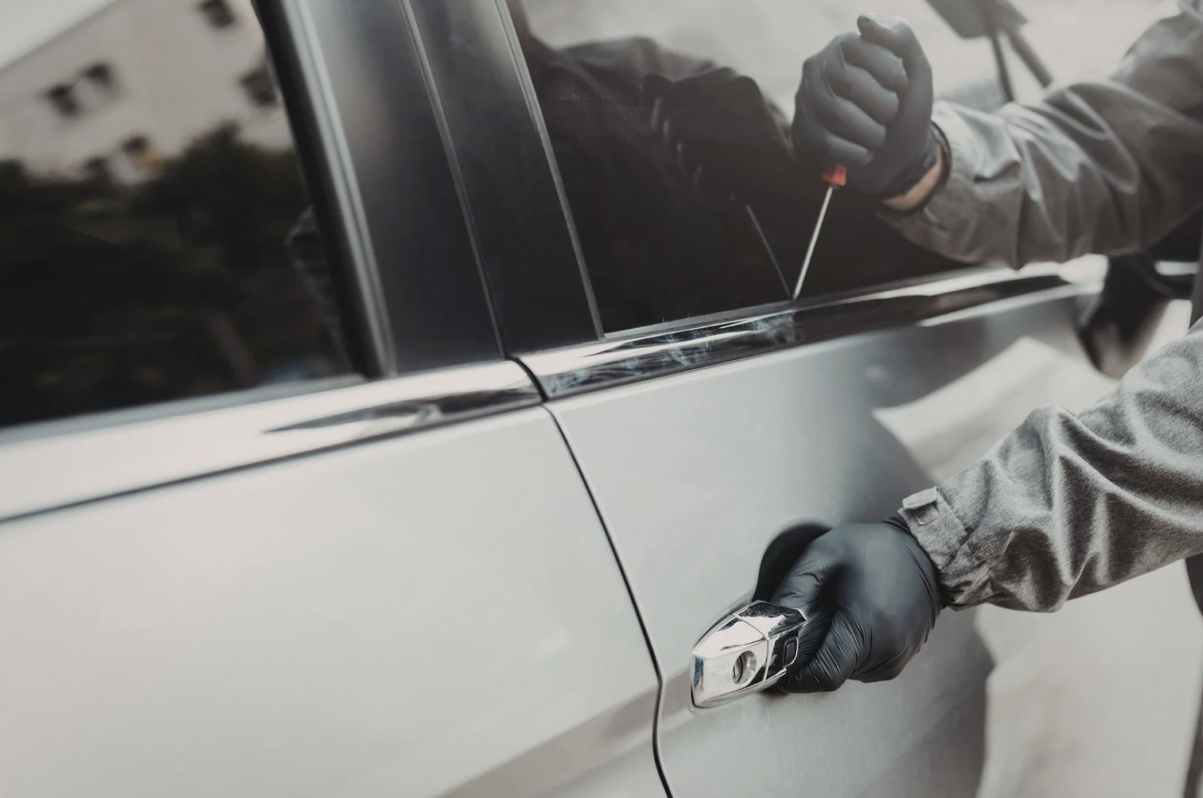 a person wearing a jacket and nylon gloves, using a screwdriver to break into a car