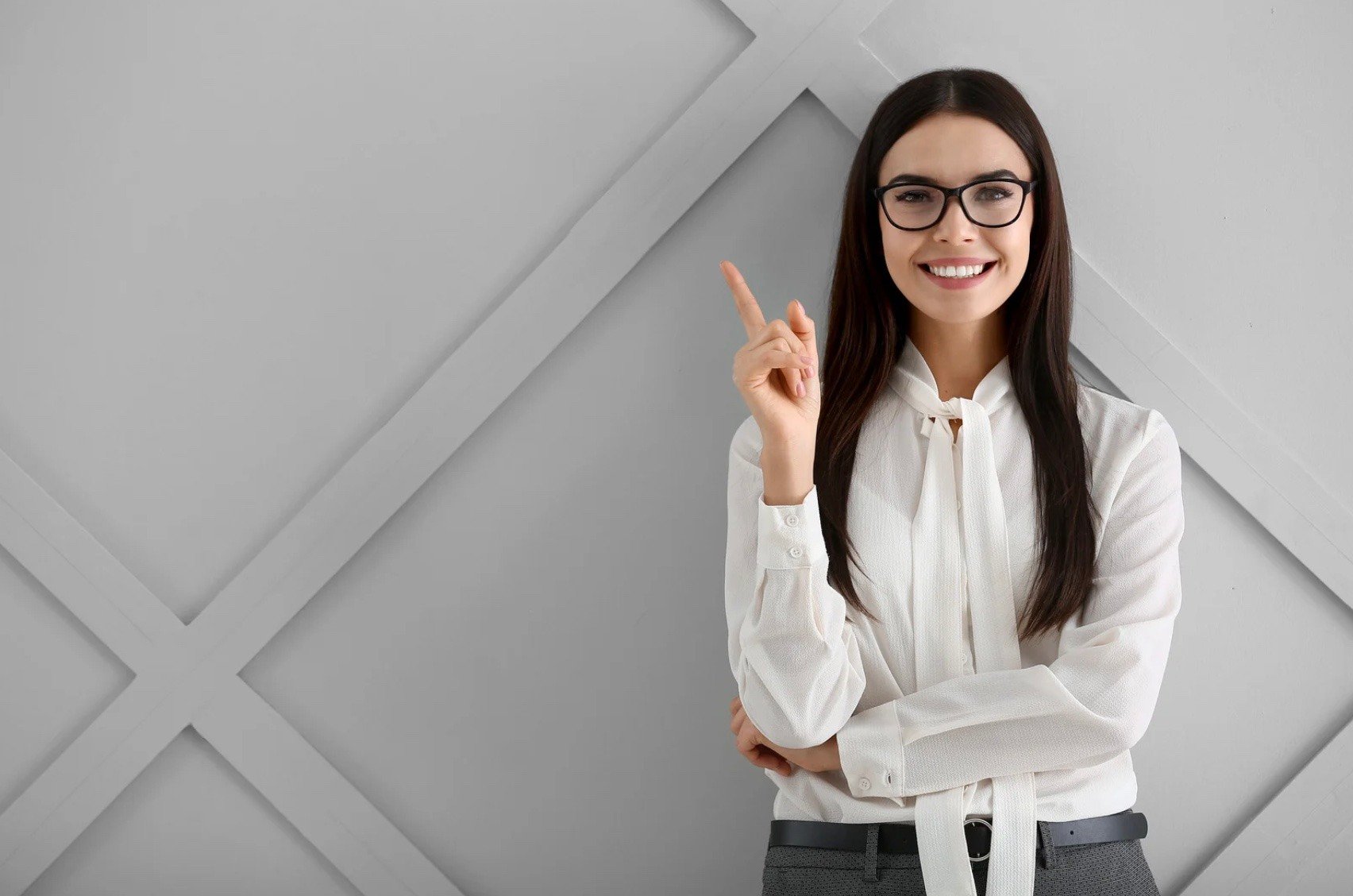 a woman wearing glasses, standing in front of a wall, pointing her finger upwards.