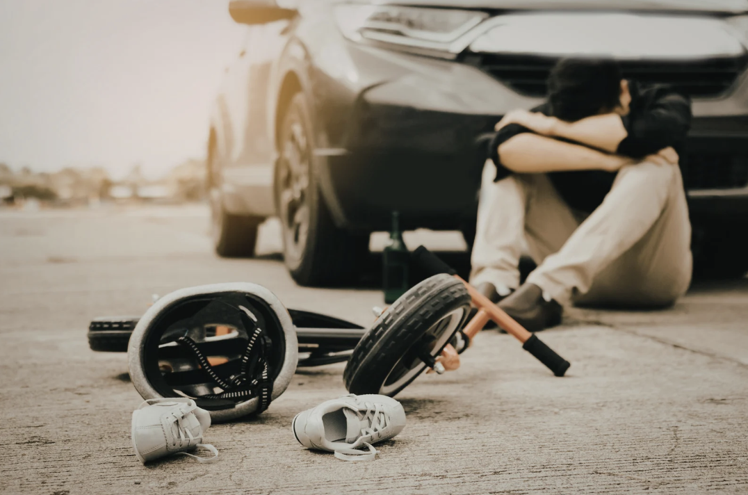a man sitting infant of his car with his head and arms on his knees and a Childs shoes and helmet damaged in front of him