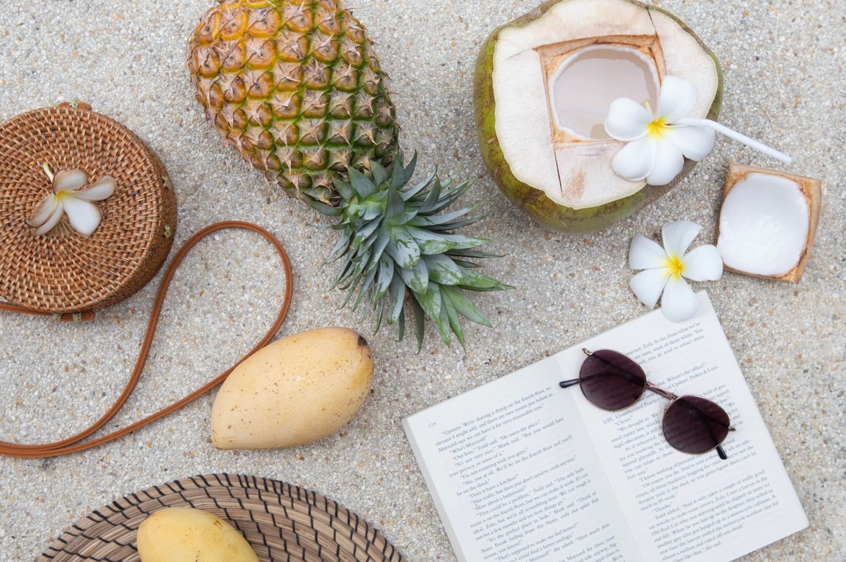 a vacation set up of a pineapple, a book, sunglasses, and a coconut